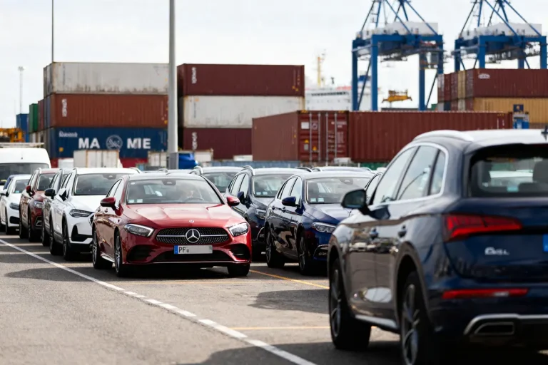 Voitures alignées devant conteneurs colorés dans un port industriel avec grues en arrière-plan.