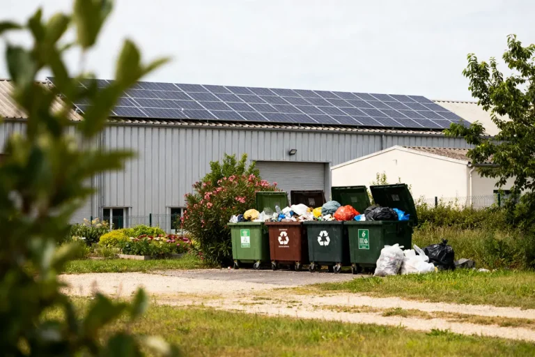 Conteneurs de recyclage débordant de déchets, devant un bâtiment avec panneaux solaires sur le toit.